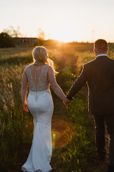 under the golden rays of sun a couple walks through a prairie field holding hands on their wedding day