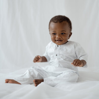 A joyful one-year-old boy sits and smiles during his early childhood milestone session. Dressed in cultural wear, he is photographed in a soft, airy studio setting filled with natural light.
