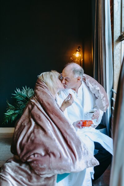 Two senior partners with gray hair are wrapped in a pink, fuzzy blanket. They are holding coffees and fruit, as they lean in and kiss each other.