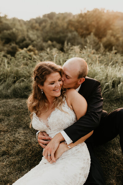 Wedding couple sitting in grass with groom's arms wrapped around the bride