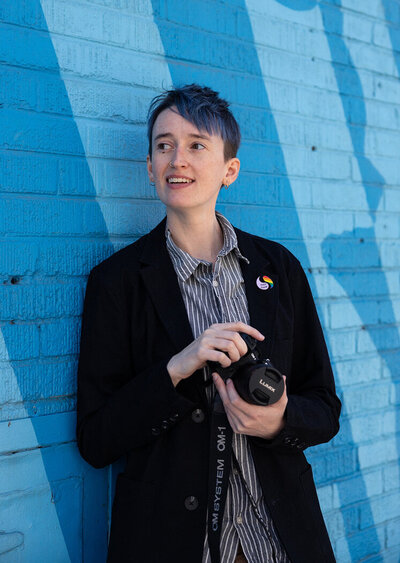 photographer River Dantzler smiles and looks off to the side, holding their camera. they have short blue hair and wear a blazer with a rainbow pride pin and "they/them" pronoun pin.