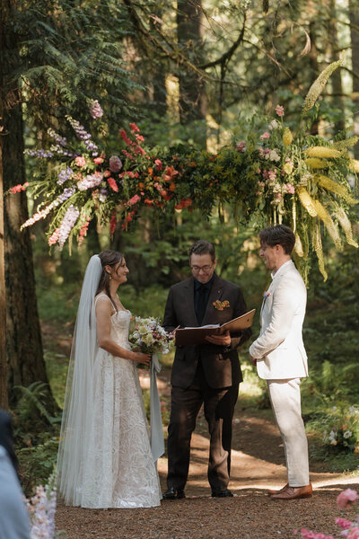 A candid photograph of a wedding ceremony in the forest at Camp Colton in Colton, Oregon. There is an extra large, colorful flower installation suspended above the couple. The flowers are bright and colorful purple, pink, and yellow. The bride is on the left and is wearing a white lace wedding dress and a floor-length veil. The groom is on the right and is wearing a light tan suit. The officiant is in the middle and is wearing a dark brown suit. Behind the people, out of focus, there is lush, green forest full of evergreen trees and ferns.