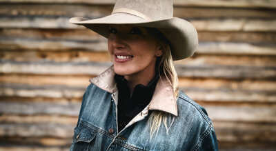 Woman stands smiling against a weathered barn in a white cowboy hat and denim jacket.