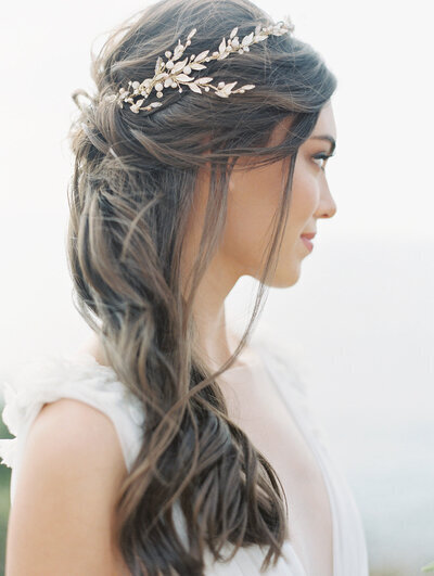Kohnur Ethereal bride with natural bronzed makeup and long loose waves styled with a gold leaf and pearl hair vine, posing on a coastal cliffside.