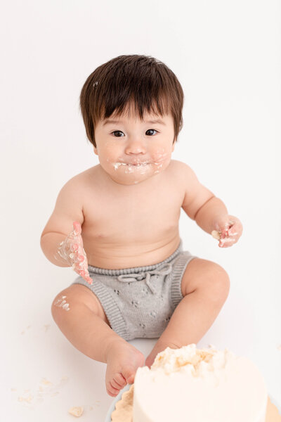 One Year old baby eating a simple, minimal birthday cake in portland baby photography studio. Baby is wearing a light grey diaper cover and has cake all over his face.