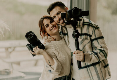 Laura et clément, souriants et enlacés, photographe et vidéaste de mariage, posent avec leurs appareils photo respectifs.