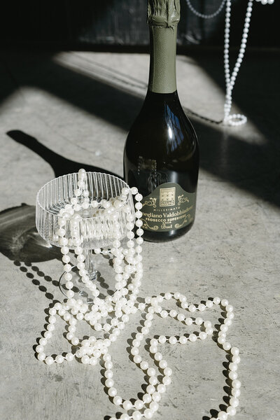 Luxurious setting with a pearl necklace in a coupe glass beside a champagne bottle at a wedding.