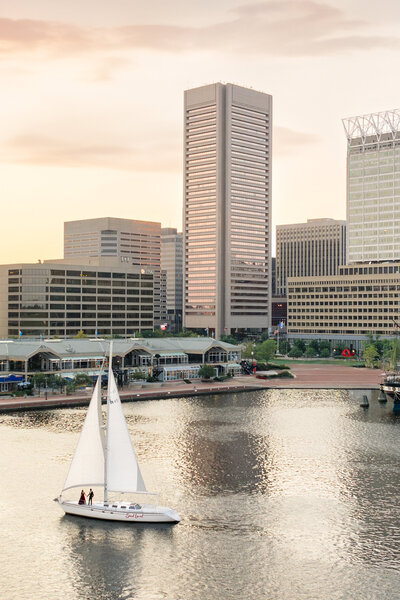 A sailboat glides on a calm river at sunset, with a citys skyline in the background. Tall buildings, including a prominent skyscraper, are bathed in warm light, reflecting on the waters surface.