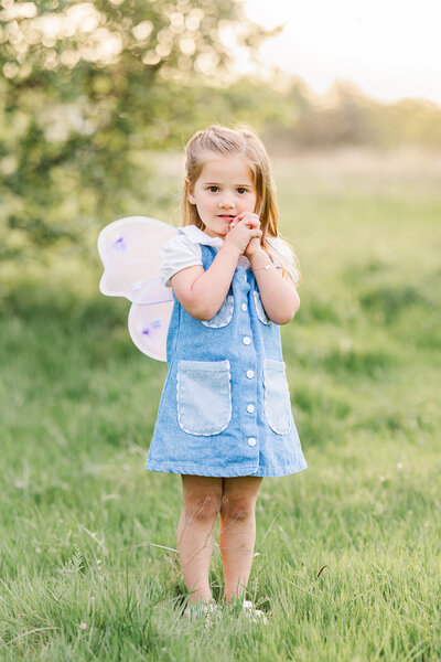 A light and airy photograph of a young girl dressed in fairy wings and a denim dress, standing in a sunlit field in the DFW area. Captured by Jennifer Lynn Photography, a Dallas Fort Worth wedding and family photographer known for timeless, natural light imagery.