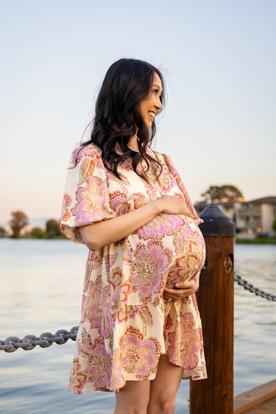 A glowing pregnant mother smiles peacefully by the water at sunset, photographed by Ellobelle Photography on a serene dockside.