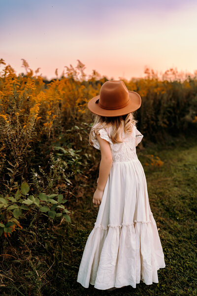 Girl wearing hat and dress looks at sunset over a field of tall grass and flowers