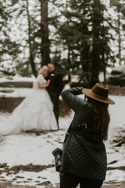 Wedding photographer in hat captures couple during snowy forest elopement in Breckenridge.