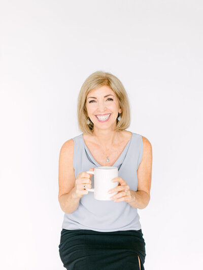 Lora Ulrich n a grey sleeveless top holding a mug, smiling at the camera