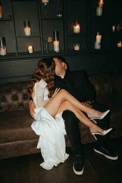 A newlywed couple shares a passionate kiss on a vintage brown leather couch in a dimly lit, moody lounge. The bride, with long cascading waves, wears a modern white wedding gown with sheer long sleeves and a thigh-high slit, draping elegantly over the couch. Her sparkling silver heels rest on her groom’s lap as she leans into him. The groom, dressed in a sleek all-black suit with casual sneakers, holds her close. Behind them, a black wall adorned with glowing candlelit shelves sets an intimate, romantic atmosphere, adding warmth and depth to the moment. Taken at Sea Cliff Manor in Long Island, NY
