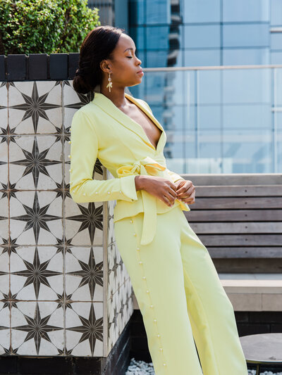 A model wearing a bright yellow pant suit leans against a wall glancing up into the midday sun . A cityscape is behind her