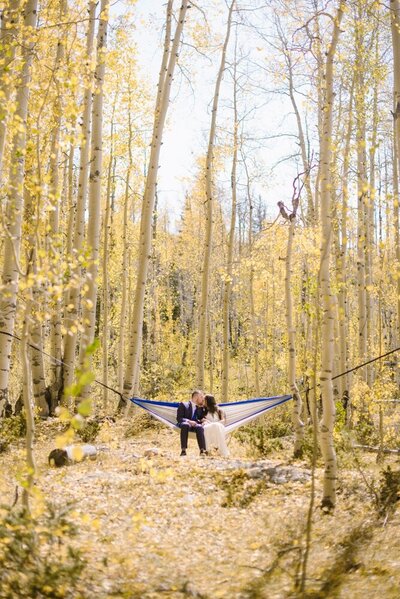 Wedding couple kisses in a white and blue hammock hanging between two aspen trees with yellow fall leaves of the trees all around.
