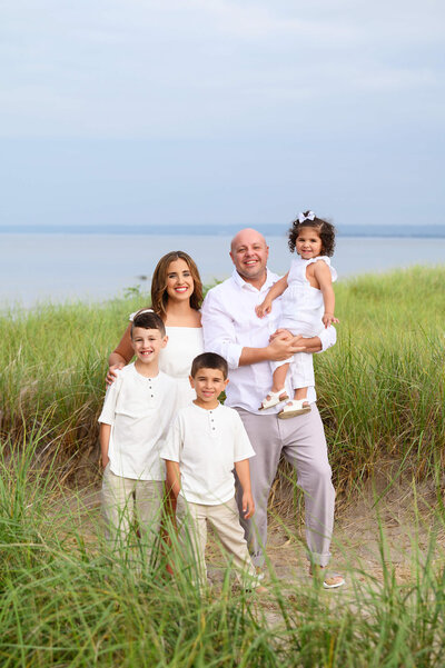 Mom and dad with their two sons and daughter standing in front of the water all in white for their beach family photography session in Burlington, Ontario.