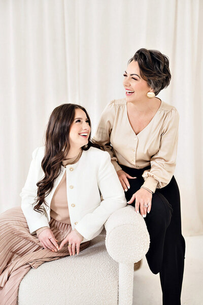 Kari and Karli Dawson seated together on a white curved sofa, smiling at each other in an intimate and joyful moment, showcasing the heart of their mother-daughter photography partnership