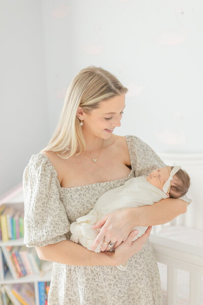 In a cozy home nursery, a mother lovingly gazes at her swaddled newborn as she cradles the baby in her arms. The warm, soft lighting creates a serene atmosphere, highlighting the tender moment between mother and child