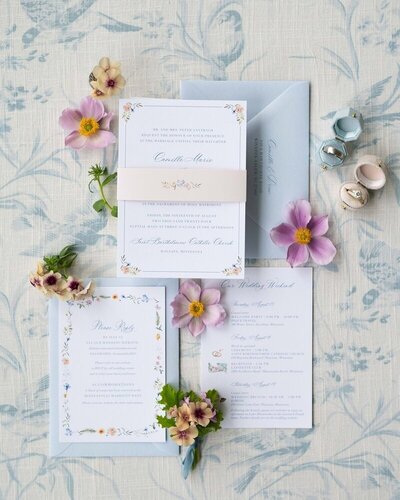 artful shot of wedding invitation details in muted pastel tones surrounded by flowers and two rings in minneapolis minnesota