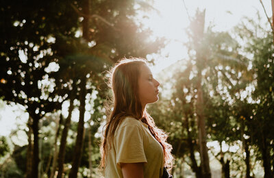 A girl standing in a forest facing sideways, her eyes are closed and she looks relaxes, the sun is shining through the trees behind her