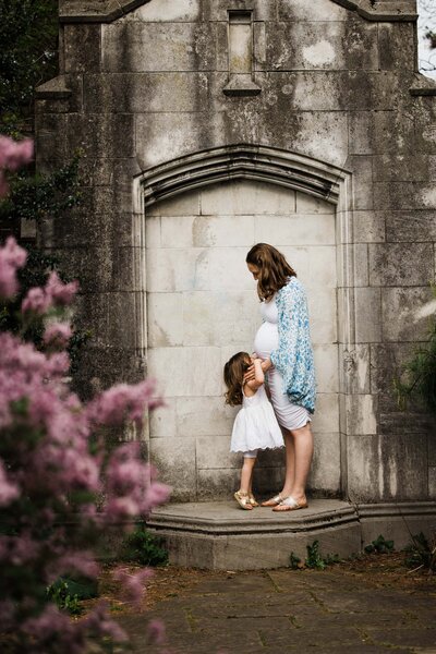 Pregnant woman and young girl standing next to each other by a stone wall, surrounded by blooms in a maternity photography session.