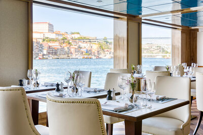 Elegant waterfront restaurant interior with neatly set tables, white upholstered chairs, and large windows overlooking a scenic river and hillside buildings.