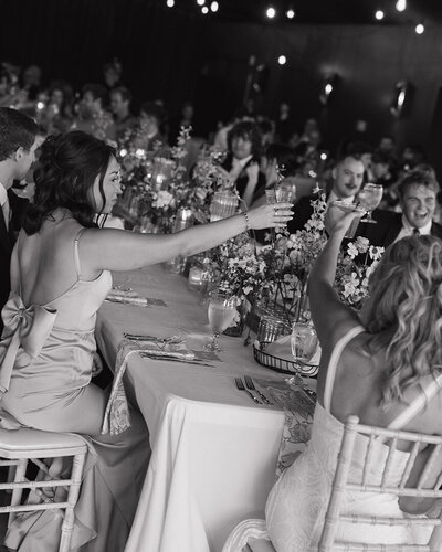 Black and white image of a joyful wedding reception. Guests seated at a floral-decorated table raise glasses for a toast, conveying celebration and warmth.