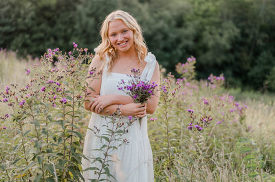 Teenage girl in white dress stands in a field with purple wildflowers.