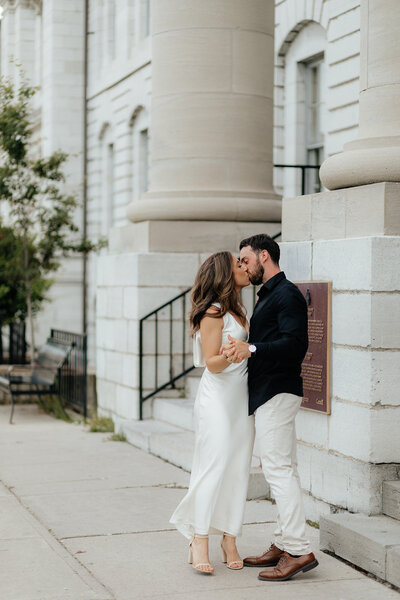 A couple kissing in front of Kingston City Hall for their engagement photoshoot.