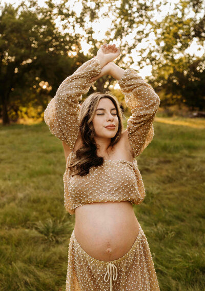Maternity mom posting in jeans and white lace crop top with belly showing. She is sitting on a boho setting at Paula Goforth's studio in Fort Worth, Texas