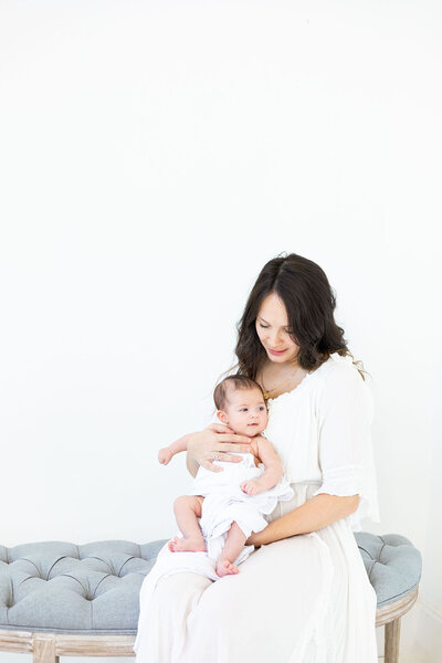 Mother holding her 2 month old baby sitting on a gray bench in a white studio NH Newborn Photographer