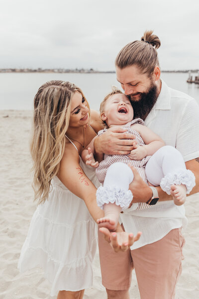 a mom and dad hold their daughter and laugh together on the beach at the Catamaran  Resort and Spa in San Diego