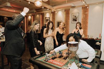 Guests cheer and laugh around a horse racing casino game table during a lively celebration at SoHo63, a Chandler wedding and event venue. Dressed in formalwear, attendees enjoy the festive atmosphere surrounded by exposed brick walls and warm lighting.