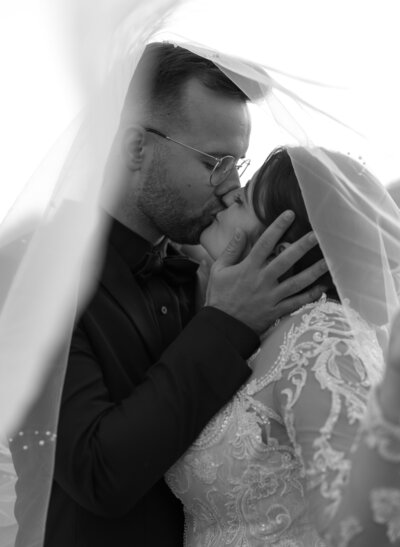 A bride and groom kiss under a veil. The groom, in glasses and a dark suit, embraces his bride in a lace wedding dress. Their hands tenderly hold each other's faces, capturing an intimate moment. This black-and-white portrait by a New England Photographer embodies fine art weddings' timeless elegance.