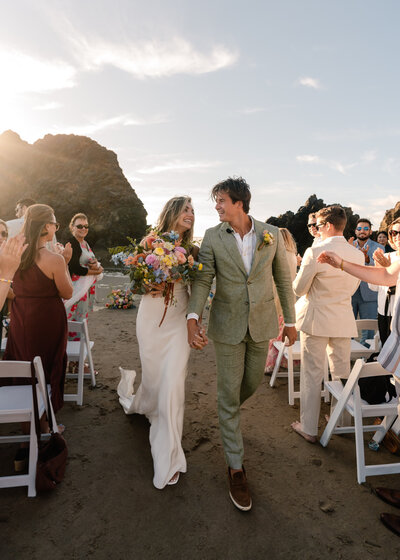 During their Oregon coast elopement, a bride and groom exit their ceremony dow a simple aisle, surrounded by their guests. they smile widely and gaze lovingly at each other.