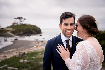 A bride, Corinne, lovingly gazes at her groom, Jordan, while he looks confidently at the camera on their wedding day by the iconic Battery Point Lighthouse in Crescent City, California.
