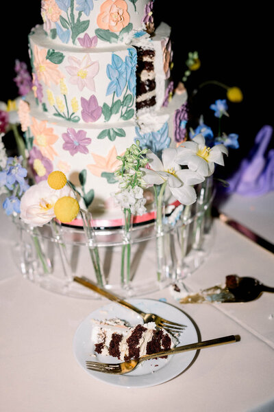 Hand-painted floral tiered wedding cake with cake slice cut out and plated on table for wedding couple.