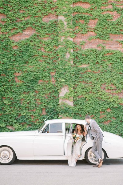 Groom helping bride out of a vintage white car in front of a red brick wall covered in green ivy, with the bride holding a bouquet and smiling.