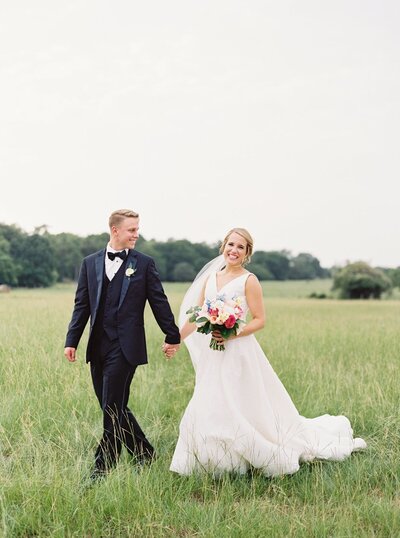Bride and groom walking in open field with blush, peach, and white bouquet