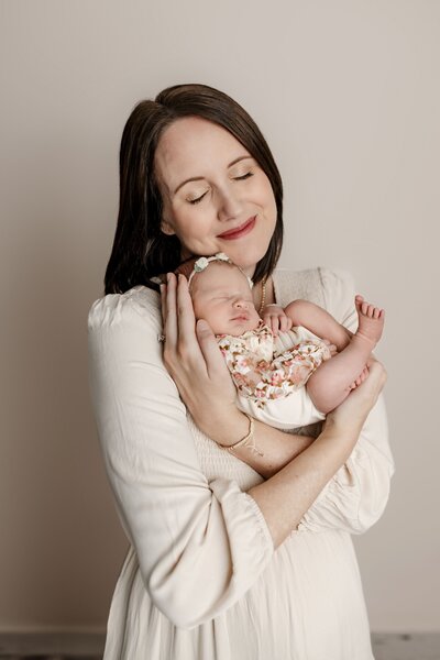 A mom in a white dress gently cuddles a newborn baby dressed in a floral outfit, against a plain background.
