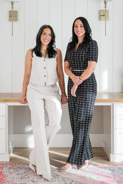 Two women in business suits stand confidently in front of a desk, representing leadership in sales and coaching.