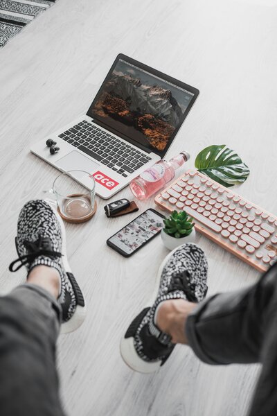 Overhead view of someone’s legs in black-and-white sneakers stretched out toward a light wood floor workspace: open laptop showing mountain wallpaper, pink retro-style keyboard, phone with floral case, small succulent, glass with iced drink, earbuds, and a monstera leaf for decoration.