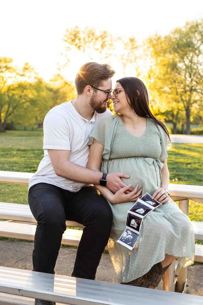 Pregnant woman with her husband sitting on bench while husband holds her baby bump and she holds a sonogram of the baby
