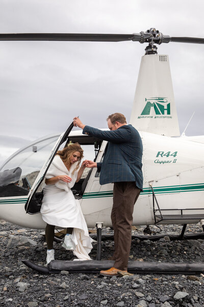 Groom holds the helicopter door so is smiling wife can exit the chopper that has just landed on glacier talus below a glacier.