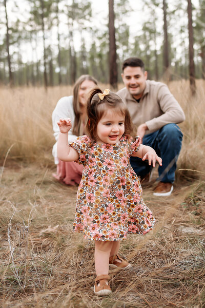 Joyful toddler in a floral dress walking toward the camera during a lifestyle family session in a pine forest near Gainesville, Florida, photographed by Salt + Heart Photography