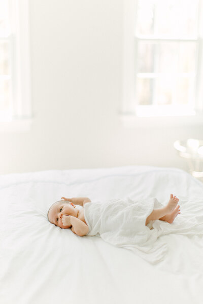 A newborn baby girl lays on a white duvet blanket with a white swaddle over her