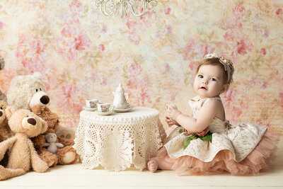 Baby girl in a gold and blush dress with a floral headband holds a tiny teacup at a lace-covered table set for tea, surrounded by plush teddy bears and delicate china, photographed by a Dallas milestone photographer.