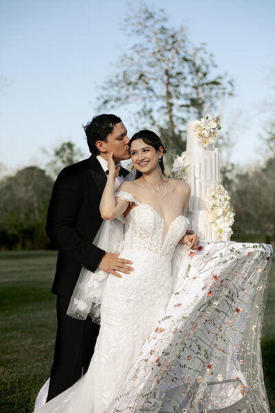Groom kissing bride on the temple next to elegant white tiered wedding cake at Maison De Veaux in Houston, Texas with embroidered veil on display
