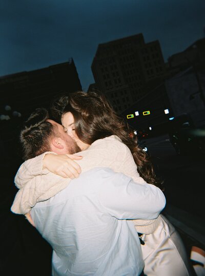 A romantic nighttime engagement photo in downtown Dallas, featuring a couple embracing on a city rooftop with urban lights glowing in the background.
